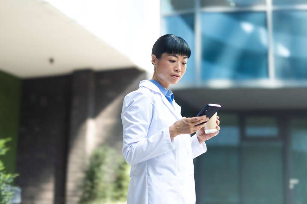 Asian female doctor checking smartphone and drinking coffee outside a modern building
