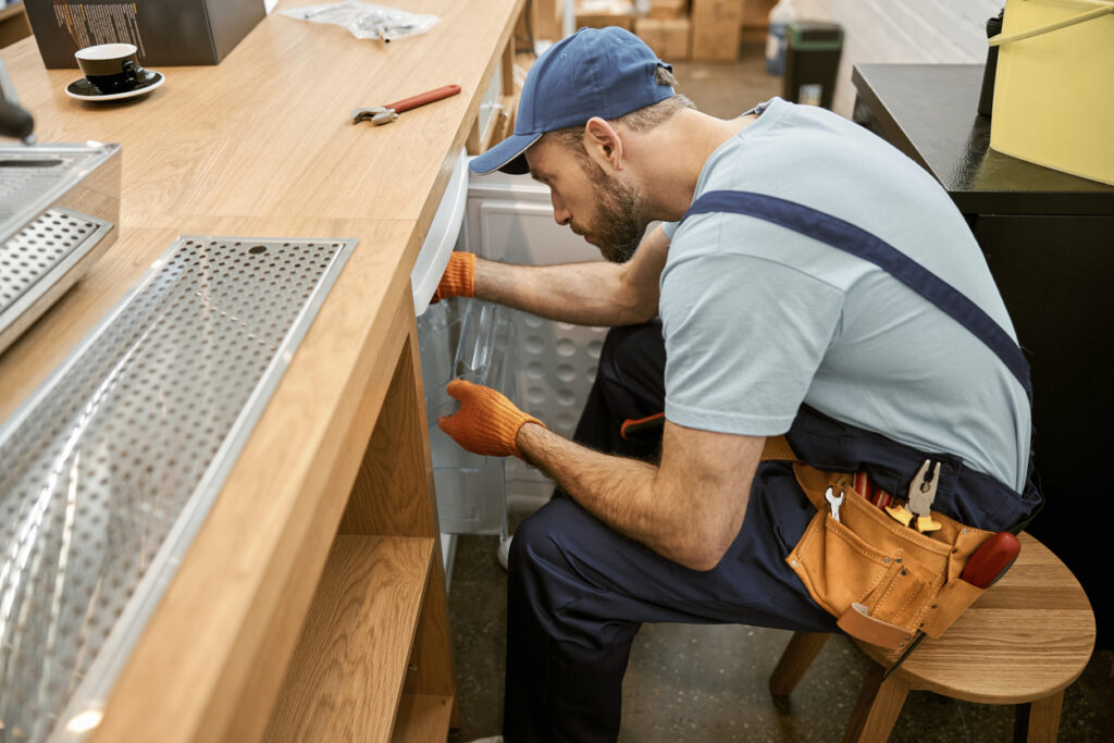 Repairman wearing work overalls with tool belt while holding plastic containers and fixing refrigerator in cafeteria