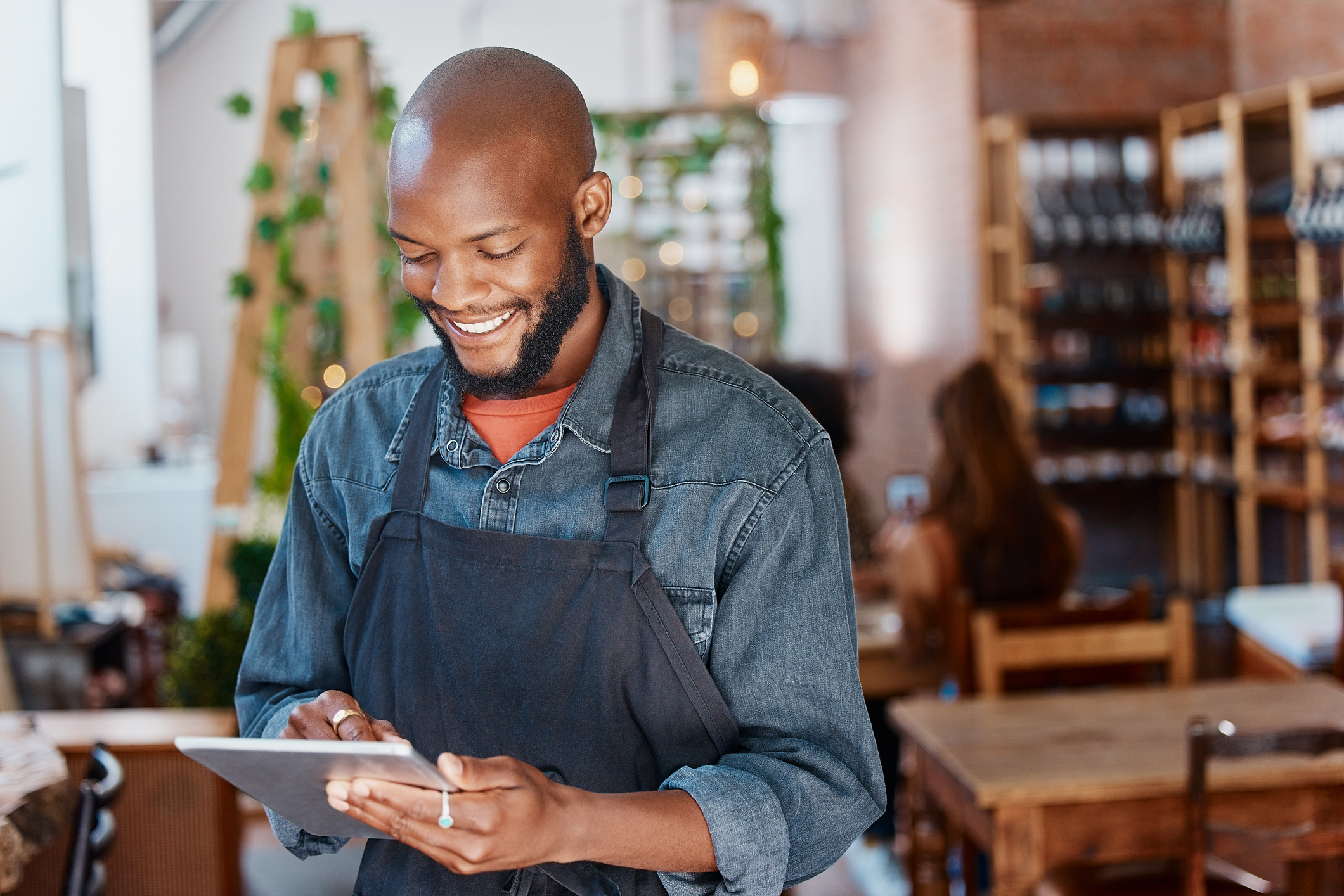Man in an apron looks at something on a tablet and smiles. He's standing in some type of cafe or wine store.