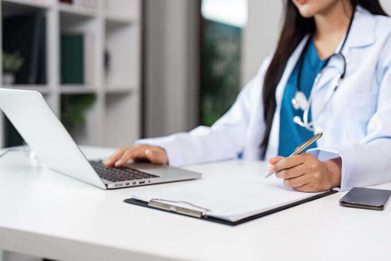 A professional doctor in a white coat working on a laptop and writing notes in a modern medical office.