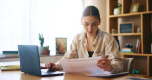 Woman, reading and document with laptop at house for loan approval, bank application and budget planning. Girl, online taxes and review paperwork for mortgage, financial invoice and investment letter MyOrders
