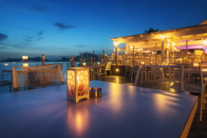 Beautiful lamp on the table in cafe on sea coast at night in summer. Landscape with candle and blurred background with chairs and tables, lights. Empty cozy cafe outdoor with illumination. Supply America patio dining supplies