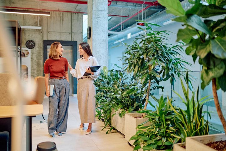Two businesswomen walking through a vibrant green office, holding tablets and engaging in a lively discussion about their work projects